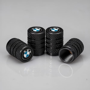 Puede incluir: Un juego de seis tapones negros para válvulas de neumáticos. Cada tapón tiene una superficie texturizada y presenta el logotipo de BMW en el centro, con un emblema azul y blanco. Los tapones están dispuestos sobre una superficie reflectante.
