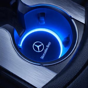 May include: A circular vehicle cup holder featuring a glowing blue LED ring light. The base of the cup holder displays the Mercedes-Benz logo and brand name in white text. The surrounding console is finished in a brushed metallic silver material.