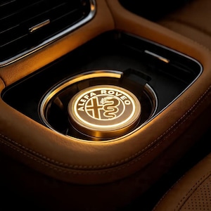 Peut inclure: Un emblème Alfa Romeo lumineux et circulaire, placé dans le porte-gobelet d'une voiture. L'emblème est doré avec un logo blanc, et l'intérieur est de couleur beige chaud. Le porte-gobelet est noir, et l'intérieur environnant est en cuir beige.