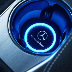 May include: A circular Mercedes-Benz branded cup holder insert featuring a glowing neon blue LED ring light. The insert is set within a brushed metallic silver console surface, displaying the Mercedes-Benz logo and text in the center.