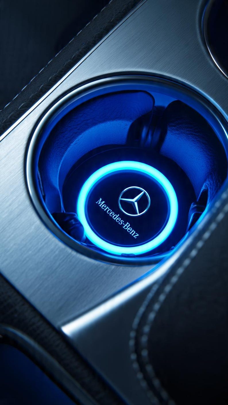 May include: A circular Mercedes-Benz branded cup holder insert featuring a glowing neon blue LED ring light. The insert is set within a brushed metallic silver console surface, displaying the Mercedes-Benz logo and text in the center.
