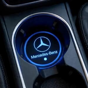 May include: A circular car cup holder insert featuring a glowing blue LED light ring. The center displays the white Mercedes-Benz logo and the text Mercedes-Benz. The insert is set within a black console with a metallic silver trim ring.