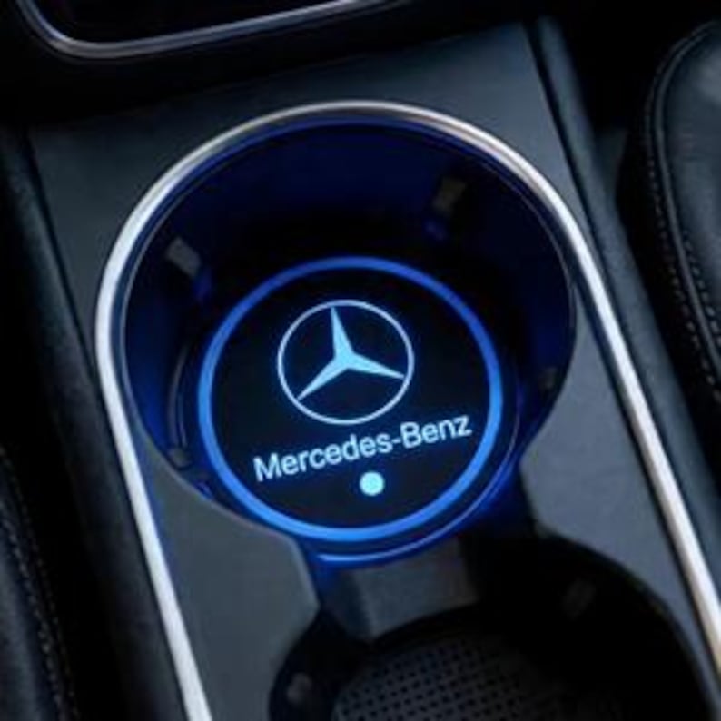 May include: A circular car cup holder insert featuring a glowing blue LED light ring. The center displays the white Mercedes-Benz logo and the text Mercedes-Benz. The insert is set within a black console with a metallic silver trim ring.