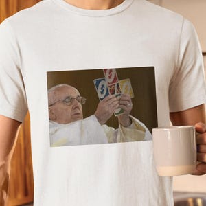 May include: White t-shirt featuring a printed image of Pope Francis holding up Uno cards. The image on the shirt shows the Pope wearing glasses and a white robe. The person in the image is also holding a beige mug.