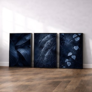 May include: Three framed botanical prints. The prints feature close-up images of leaves and plants in shades of blue and dark navy. The frames are black, and the prints are displayed on a wooden floor against a white wall.