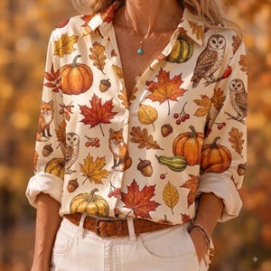 May include: A long-sleeve button-down shirt in a cream color featuring an all-over print of orange pumpkins, brown acorns, red berries, yellow and red maple leaves, and brown owls and foxes. The shirt is tucked into white pants with a brown belt.
