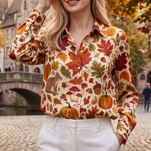 Könnte beinhalten: Ein langärmeliges Hemd mit Knopfleiste auf cremefarbenem Grund und einem herbstlichen Druckmuster. Das Design zeigt orangefarbene und rote Ahornblätter, braune Eicheln, rot-weiße Pilze, orangefarbene Kürbisse und ein kleines braunes Eichhörnchen.