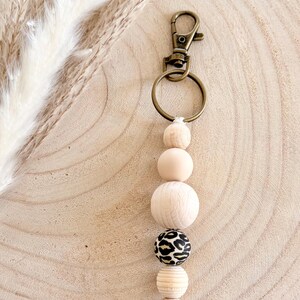 May include: A beaded keychain with a gold-colored clasp and ring. The keychain features a series of wooden beads in various shapes and sizes, including a bead with a black and tan leopard print. The beads are strung together on a white cord.
