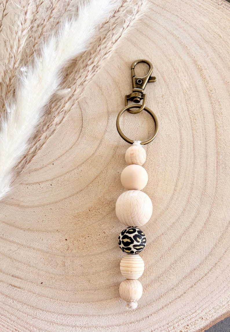 May include: A beaded keychain with a gold-colored clasp and ring. The keychain features a series of wooden beads in various shapes and sizes, including a bead with a black and tan leopard print. The beads are strung together on a white cord.