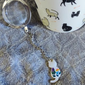 Op de afbeelding: Een witte keramische mok met zwarte kattenillustraties en een roestvrijstalen thee-ei. De infuser heeft een ketting en een witte kattenhanger met kleurrijke bloemenaccenten. De mok is gevuld met thee.