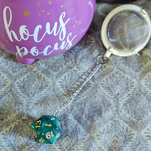 Könnte beinhalten: Eine lila Keramiktasse mit dem Schriftzug "hocus pocus" in weißer Schrift, akzentuiert mit goldenen Sternen. Ein silberner Tee-Ei mit einer Kette ist an einem teal- und goldfarbenen Polyederwürfel befestigt.
