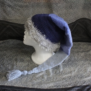 Könnte beinhalten: Eine blaue und hellblaue Strickmütze mit langem Schwanz und weißem Pelzbesatz. Die Mütze sitzt auf einem Mannequin-Kopf.