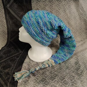 Op de afbeelding: Een blauwe, groene en witte gestreepte gebreide muts met een lange staart. De muts zit op een wit mannequin hoofd.