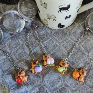 Op de afbeelding: Theezeefjes met berenhangers met fruit, waaronder een aardbei, een pruim, een peer en een sinaasappel. Een witte mok met zwarte kattenillustraties en roestvrijstalen thee-infusers zijn ook aanwezig.