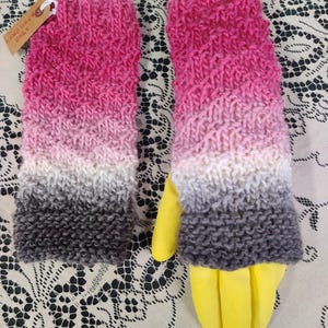 Könnte beinhalten: Ein Paar handgestrickte Fingerhandschuhe in einem Farbverlauf aus Rosa, Weiß und Grau. Die Handschuhe haben ein strukturiertes Strickmuster und ein Etikett mit der Aufschrift "HAND WASH ONLY".