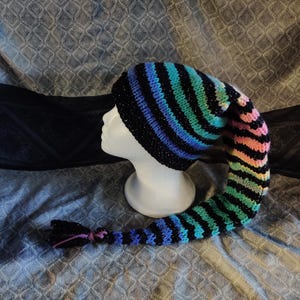 May include: A colourful, striped knitted hat with a long, trailing tail. The hat features black, blue, green, and pink stripes. The tail is finished with a tassel. The hat is displayed on a white mannequin head.