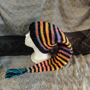 May include: A brightly coloured knitted hat with a long, pointed tail and a tassel. The hat has vertical stripes in black, orange, yellow, pink, and blue. The tassel is black and blue. The hat is displayed on a white mannequin head.