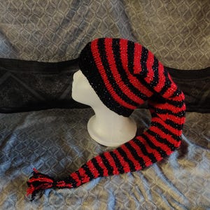 Op de afbeelding: Een rood-zwarte gestreepte gebreide muts met een lange, puntige staart. De hoed wordt getoond op een witte mannequinhoofd. De staart eindigt in een kwast. De hoed is waarschijnlijk handgemaakt.