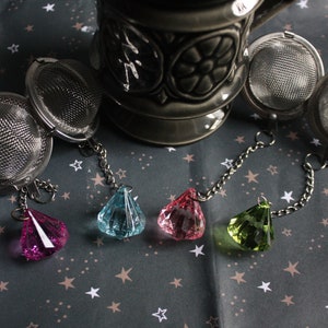 May include: Four silver tea infusers with mesh balls and chains, each with a different coloured crystal charm: pink, blue, green, and clear.