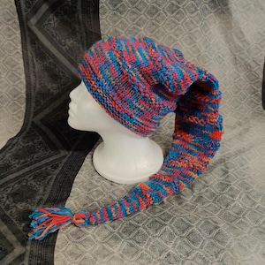 Op de afbeelding: Een kleurrijke gebreide muts met een lange staart. De muts is blauw, roze en oranje met een witte garenkwast aan het einde van de staart.