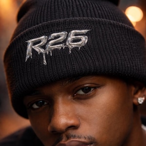 May include: Black knit beanie with the silver text "R26" in a dripping style. The beanie is worn by a person with dark skin and a close-up shot focuses on the hat and face.