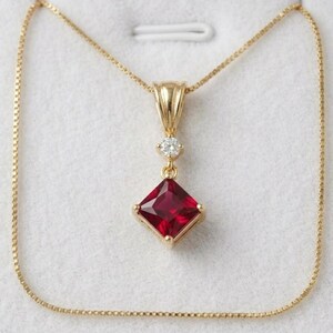 May include: A gold necklace with a square, deep red gemstone pendant. The pendant is framed in gold and has a small, round diamond above the red stone. The chain is a delicate gold link.