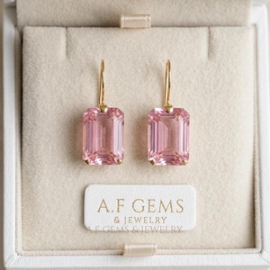 May include: A pair of gold drop earrings featuring rectangular, emerald-cut pink gemstones. The earrings are displayed in a beige jewelry box with a white label printed with the text A.F GEMS & JEWELRY. The design includes simple gold hooks.