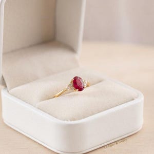 Puede incluir: Un anillo de oro con un rubí rojo de talla ovalada en el centro, flanqueado por pequeños diamantes transparentes a cada lado. El anillo se presenta dentro de un joyero blanco con un forro interior de terciopelo color crema y exterior liso.