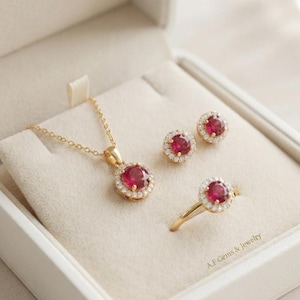 Puede incluir: Un conjunto de joyería que incluye un collar de oro con un colgante de piedra roja redonda, pendientes de botón de piedra roja redonda a juego y un anillo de oro con una piedra roja redonda, rodeados de pequeñas piedras blancas, con texto A.F Gems & Jewelry.