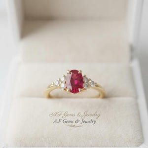 Op de afbeelding: Een gouden ring met een centrale ovaal geslepen rode robijn, geflankeerd door drie kleine rond geslepen witte diamanten aan elke kant. De tekst A.F Gems & Jewelry staat onder de ring in een lichtgekleurd doosje met een zachte textuur.
