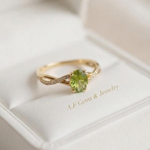 Könnte beinhalten: Ein Goldring mit einem oval geschliffenen, hellgrünen Edelstein in der Mitte, flankiert von kleinen, klaren Steinen entlang der geteilten Ringschiene. Der Text A.F Gems & Jewelry ist in Gold auf der weißen Oberfläche unter dem Ring gedruckt.