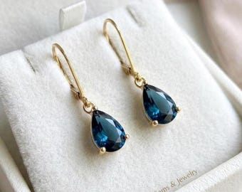 May include: A pair of gold drop earrings featuring faceted, pear-shaped dark blue gemstones suspended from thin gold hooks. The earrings are displayed on a soft, light-colored fabric surface with the text A.F Gems & Jewelry printed in the corner.