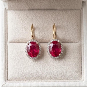May include: A pair of gold leverback earrings, each featuring a large, oval-cut, deep red ruby gemstone surrounded by a halo of small, clear, round-cut diamonds. The earrings are displayed symmetrically, showcasing the vibrant red stones and the metallic gold settings.
