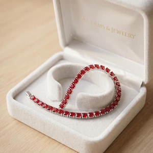 May include: A silver-toned tennis bracelet featuring a continuous row of square-cut, vibrant red gemstones. The bracelet is displayed inside an open, light-colored, velvet-lined jewelry box with the text AJ GEMS & JEWELRY printed in gold.