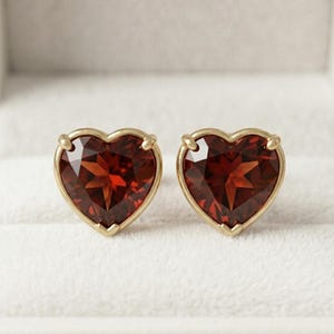 Peut inclure: Une paire de boucles d'oreilles à clous avec des pierres précieuses grenat rouge foncé en forme de cœur à facettes, serties dans des montures en métal doré poli. Chaque boucle est fixée par trois petites griffes maintenant la pierre.