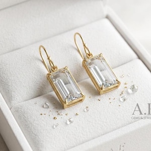 May include: A pair of gold drop earrings featuring rectangular baguette-cut clear gemstones set in polished gold frames with curved ear wires. The earrings are displayed alongside small loose clear crystals on a soft white velvet surface.