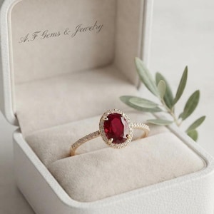 Könnte beinhalten: Ein Ring mit einem oval geschliffenen roten Rubin, gefasst in ein Goldband mit einem Kranz aus kleinen weißen Diamanten. Der Ring befindet sich in einer hellen Schmuckschatulle, auf deren Innendeckel der Text A.F Gems & Jewelry schwarz gedruckt ist.