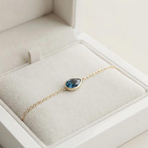 Puede incluir: Una delicada pulsera de cadena dorada con una sola piedra preciosa azul en forma de pera engastada en un bisel de oro. La pulsera se muestra dentro de un joyero de color claro forrado en terciopelo, resaltando el contraste entre el metal y la piedra.