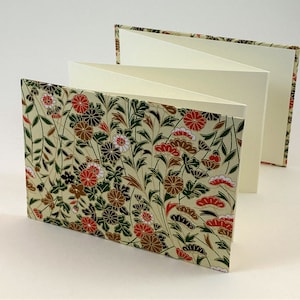 May include: A cream-colored, accordion-style book with a floral pattern cover. The cover features red, brown, and black flowers on a cream background.
