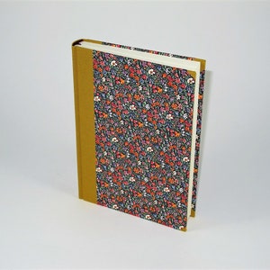May include: A hardcover book with a floral pattern cover and a mustard-colored spine. The cover features a dark background with a repeating pattern of small red, orange, and white flowers. The book is standing upright.