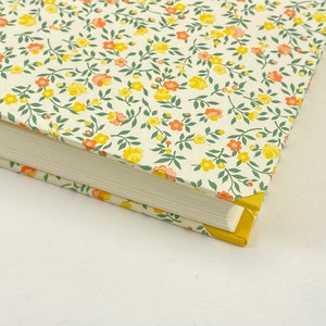 Puede incluir: Un libro blanco con una cubierta de patr&oacute;n floral. La cubierta presenta peque&ntilde;as flores amarillas y naranjas con hojas verdes sobre un fondo blanco. El libro tiene un borde amarillo.