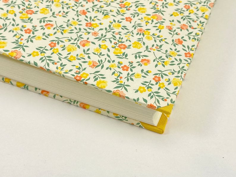 Puede incluir: Un libro blanco con una cubierta de patr&oacute;n floral. La cubierta presenta peque&ntilde;as flores amarillas y naranjas con hojas verdes sobre un fondo blanco. El libro tiene un borde amarillo.