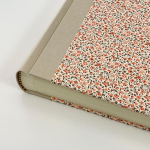 May include: A hardcover book with a beige linen spine and a floral patterned cover. The cover features a cream background with small red and brown flowers and green leaves. The book's pages are visible.