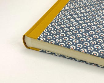 May include: A hardcover book featuring a mustard yellow fabric spine and a dark blue cover with a repeating pattern of white and orange stylized circular motifs. The book is shown from a high angle, highlighting the bound edge and paper pages.