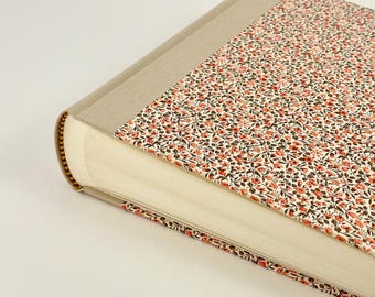 Könnte beinhalten: Ein cremefarbenes Fotoalbum mit einem floralen Muster auf dem Einband. Der Einband besteht aus einem Stoff mit einem kleinen, sich wiederholenden Muster aus roten und braunen Blumen auf weißem Hintergrund.