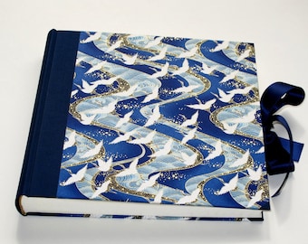 May include: A navy blue photo album with a white and gold crane pattern on the cover. The album has a blue ribbon tied around it.