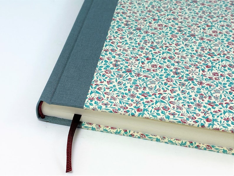 Puede incluir: Un diario de tapa dura azul turquesa con un patr&oacute;n floral en la portada. El diario tiene un marcap&aacute;ginas de cinta color burdeos.