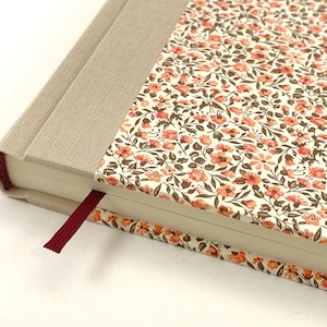 Könnte beinhalten: Ein Hardcover-Notizbuch mit beigem Leinenrücken und floral gemustertem Einband. Der Einband hat einen cremefarbenen Hintergrund mit kleinen orangefarbenen und braunen Blumen. Ein burgunderfarbenes Lesezeichenband ist sichtbar.