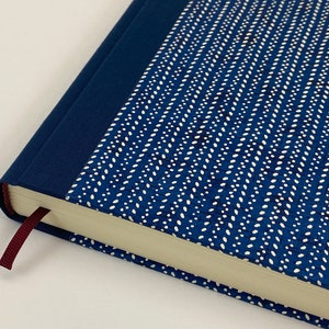 May include: A blue hardcover journal with a white patterned design and a burgundy ribbon bookmark. The journal has a blue spine and a white page edge.