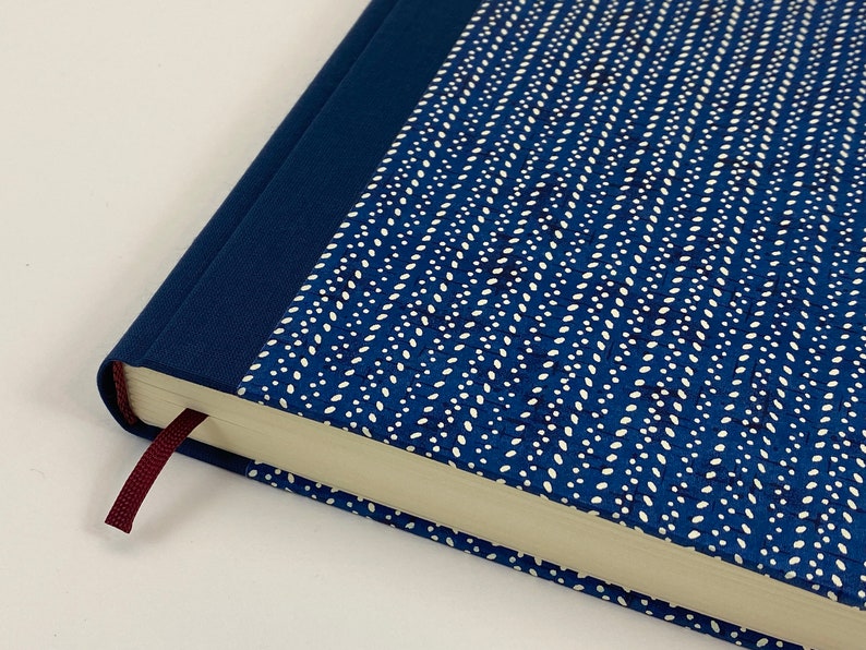 Pode incluir: Um di&aacute;rio de capa dura azul com um design estampado branco e um marcador de fita cor vinho. O di&aacute;rio tem uma lombada azul e uma borda de p&aacute;gina branca.